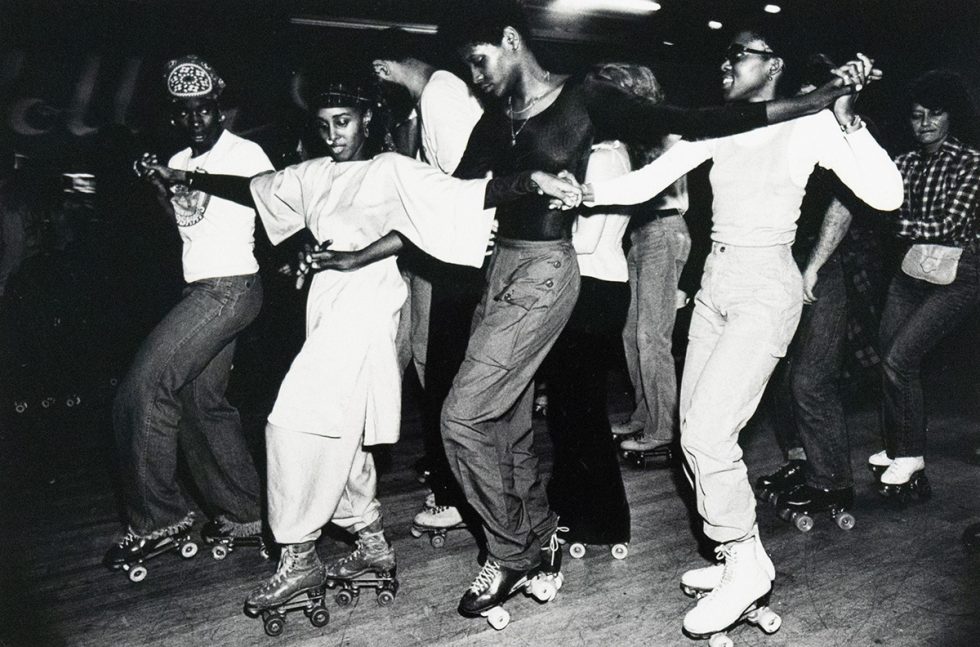 A group of roller skaters, predominantly Black, are captured in a black and white photograph performing a line dance at an indoor rink. They are holding hands and extending their arms, moving in unison across the frame.