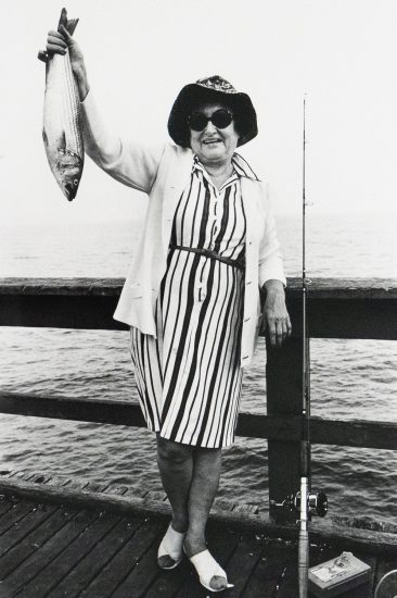 A smiling woman wearing a striped dress, light cardigan, and sun hat holds up a fish in one hand, standing on a pier beside a fishing rod. A body of water is visible behind her.
