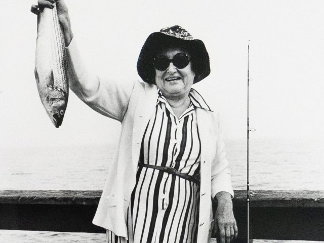 A smiling woman wearing a striped dress, light cardigan, and sun hat holds up a fish in one hand, standing on a pier beside a fishing rod. A body of water is visible behind her.