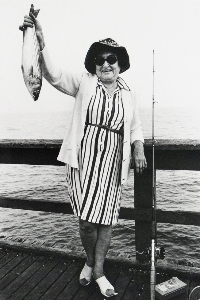 A smiling woman wearing a striped dress, light cardigan, and sun hat holds up a fish in one hand, standing on a pier beside a fishing rod. A body of water is visible behind her.