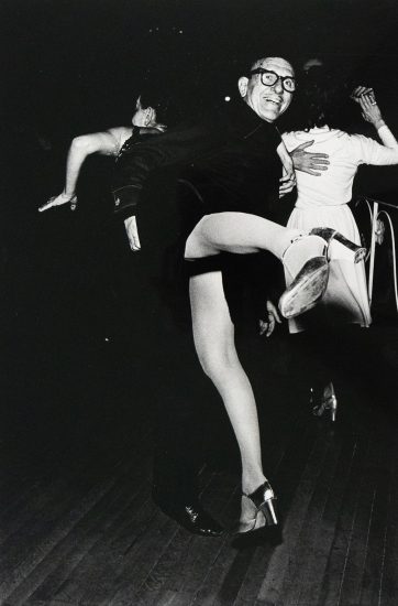 A black and white photograph captures three dancers in motion on a dark dance floor. One man in glasses smiles at the viewer while a woman's leg is dramatically extended towards the foreground.