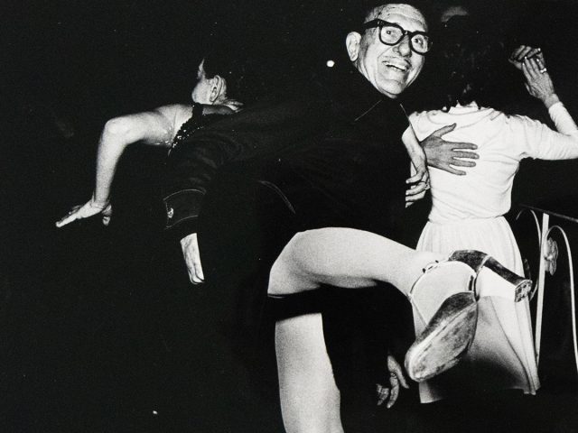 A black and white photograph captures three dancers in motion on a dark dance floor. One man in glasses smiles at the viewer while a woman's leg is dramatically extended towards the foreground.
