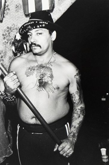 A black and white photograph captures a shirtless, tattooed man wearing a fedora, holding a stick, and a bird perched on his shoulder. He gazes directly at the viewer, his chest and arms adorned with intricate tattoo designs.