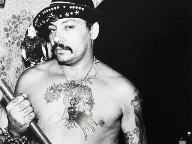 A black and white photograph captures a shirtless, tattooed man wearing a fedora, holding a stick, and a bird perched on his shoulder. He gazes directly at the viewer, his chest and arms adorned with intricate tattoo designs.