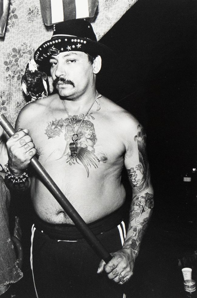A black and white photograph captures a shirtless, tattooed man wearing a fedora, holding a stick, and a bird perched on his shoulder. He gazes directly at the viewer, his chest and arms adorned with intricate tattoo designs.