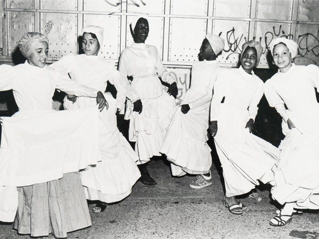 Black-and-white photograph of six young women in white dresses and head coverings dancing on a city street at night. They are mid-motion, smiling or focused, with a graffitied metal wall in the background.
