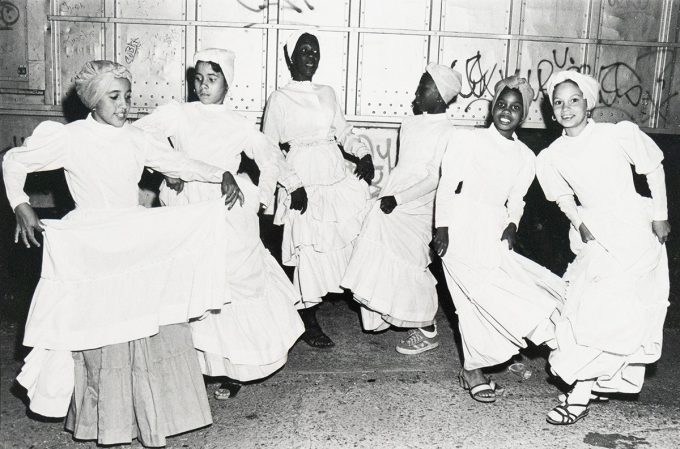 Black-and-white photograph of six young women in white dresses and head coverings dancing on a city street at night. They are mid-motion, smiling or focused, with a graffitied metal wall in the background.