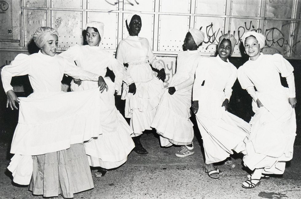 Black-and-white photograph of six young women in white dresses and head coverings dancing on a city street at night. They are mid-motion, smiling or focused, with a graffitied metal wall in the background.