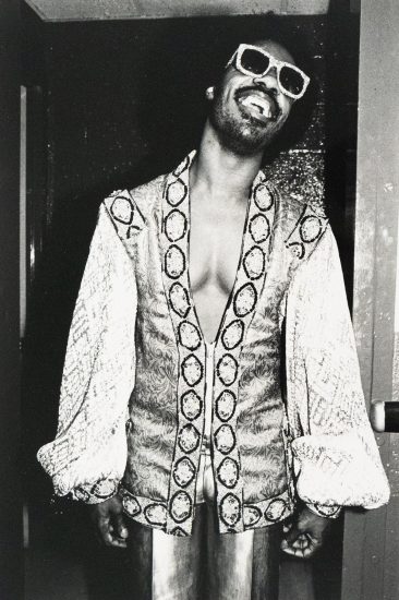 A Black man with a mustache and goatee, identified as Stevie Wonder, smiles broadly while wearing large, light-colored sunglasses and an ornate, open-front jacket. The black and white photograph captures him indoors against a dark doorway.