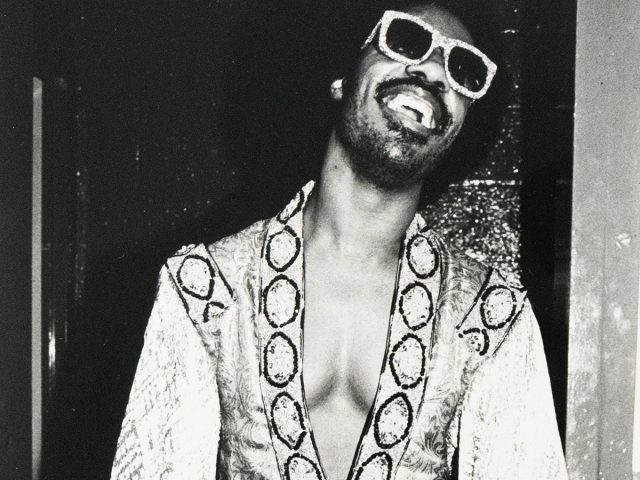 A Black man with a mustache and goatee, identified as Stevie Wonder, smiles broadly while wearing large, light-colored sunglasses and an ornate, open-front jacket. The black and white photograph captures him indoors against a dark doorway.