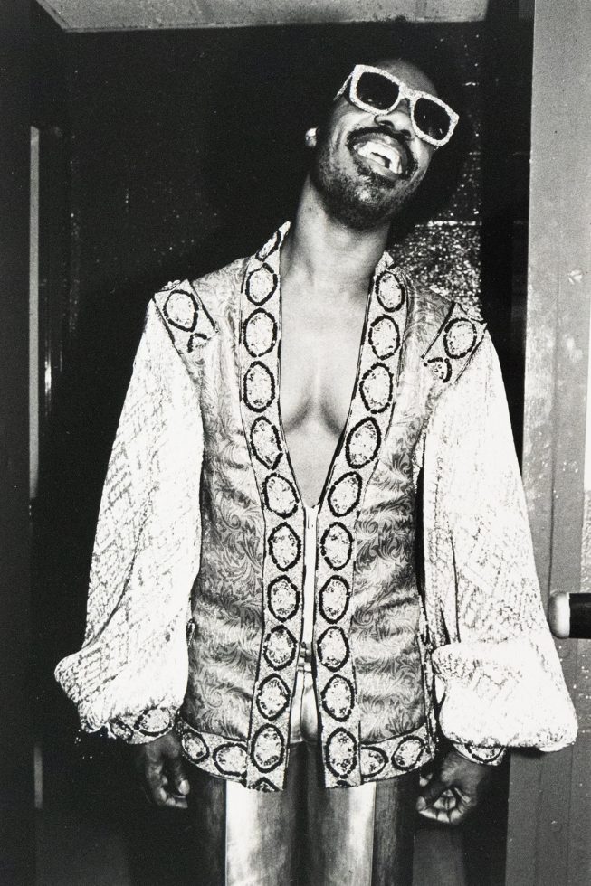A Black man with a mustache and goatee, identified as Stevie Wonder, smiles broadly while wearing large, light-colored sunglasses and an ornate, open-front jacket. The black and white photograph captures him indoors against a dark doorway.