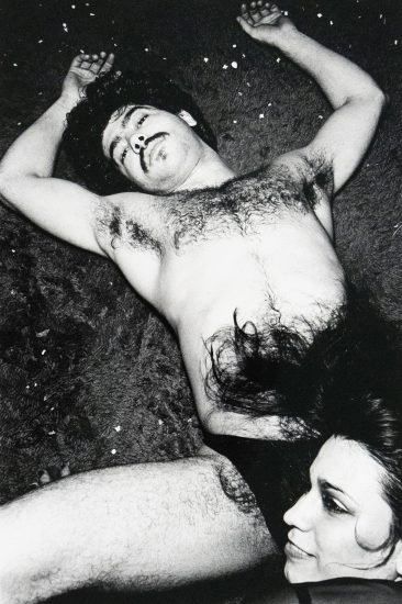 Black-and-white photograph of a shirtless man lying on a carpeted floor with his arms outstretched, looking toward the camera. His torso and facial hair are prominent. A woman's face and hair appear in the lower right corner, partially obscured by his leg. Small pieces of debris are scattered across the carpet.