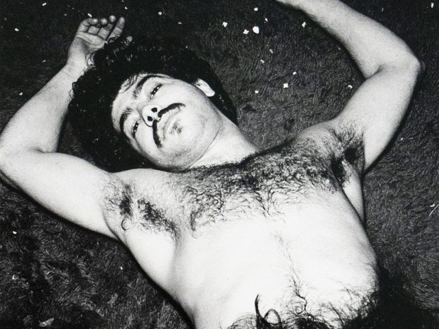 Black-and-white photograph of a shirtless man lying on a carpeted floor with his arms outstretched, looking toward the camera. His torso and facial hair are prominent. A woman's face and hair appear in the lower right corner, partially obscured by his leg. Small pieces of debris are scattered across the carpet.