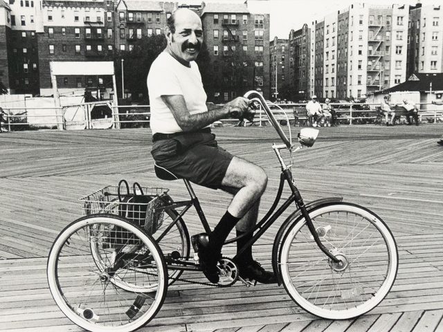 A black and white photograph captures a man with a mustache on a tricycle, smiling directly at the viewer on a boardwalk. In the background, buildings line the horizon, and other people are visible on the boardwalk.