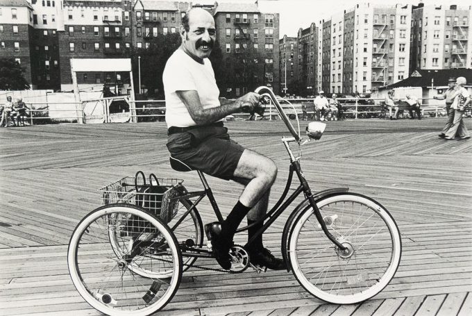 A black and white photograph captures a man with a mustache on a tricycle, smiling directly at the viewer on a boardwalk. In the background, buildings line the horizon, and other people are visible on the boardwalk.