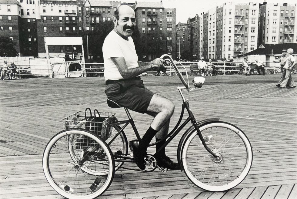 A black and white photograph captures a man with a mustache on a tricycle, smiling directly at the viewer on a boardwalk. In the background, buildings line the horizon, and other people are visible on the boardwalk.