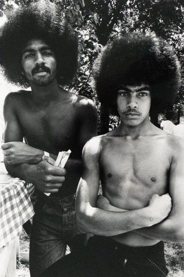 Two shirtless men with large Afros stand outdoors, facing the viewer. One man, on the left, holds what appears to be paper currency, while the other stands with crossed arms.