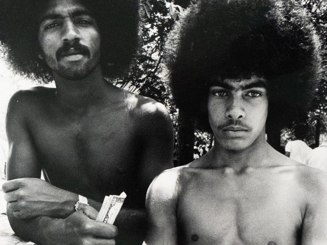 Two shirtless men with large Afros stand outdoors, facing the viewer. One man, on the left, holds what appears to be paper currency, while the other stands with crossed arms.