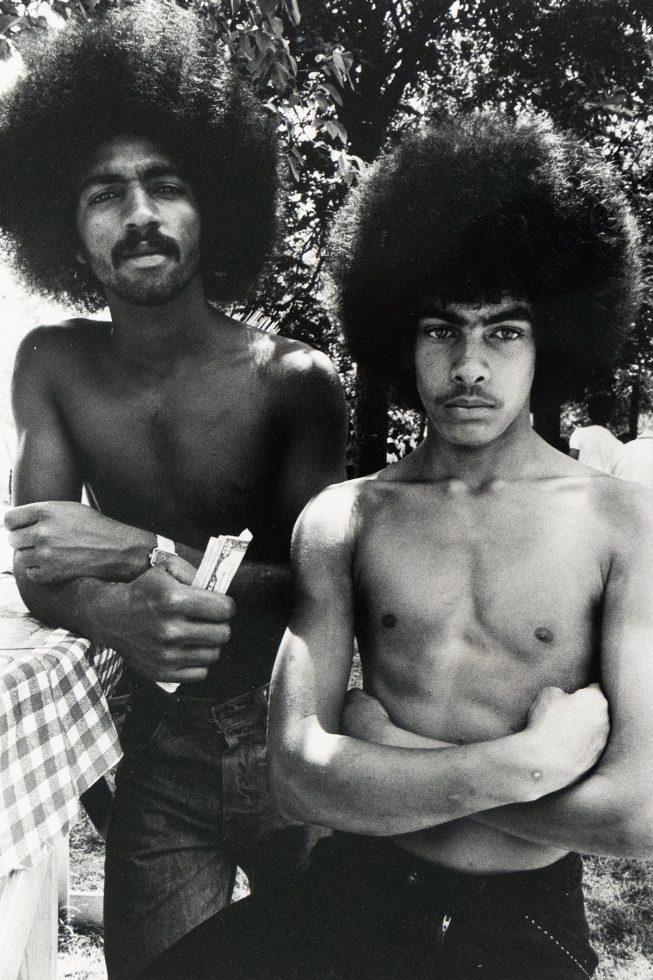 Two shirtless men with large Afros stand outdoors, facing the viewer. One man, on the left, holds what appears to be paper currency, while the other stands with crossed arms.