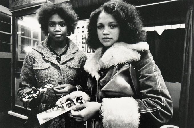 A black and white photograph captures two women standing side-by-side, each holding a strip of photographs. The woman on the left wears a textured coat, and the woman on the right wears a coat with a fur collar and cuffs, holding a clutch bag.