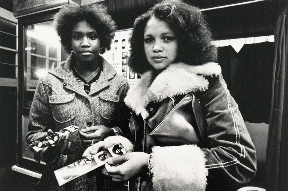 A black and white photograph captures two women standing side-by-side, each holding a strip of photographs. The woman on the left wears a textured coat, and the woman on the right wears a coat with a fur collar and cuffs, holding a clutch bag.