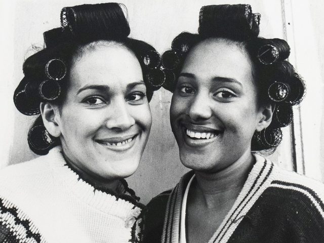 A black and white close-up captures two smiling women with dark hair in curlers, facing forward. The woman on the left wears a light-colored, patterned sweater, while the woman on the right wears a striped sweater.