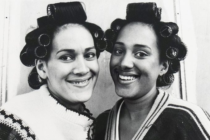 A black and white close-up captures two smiling women with dark hair in curlers, facing forward. The woman on the left wears a light-colored, patterned sweater, while the woman on the right wears a striped sweater.