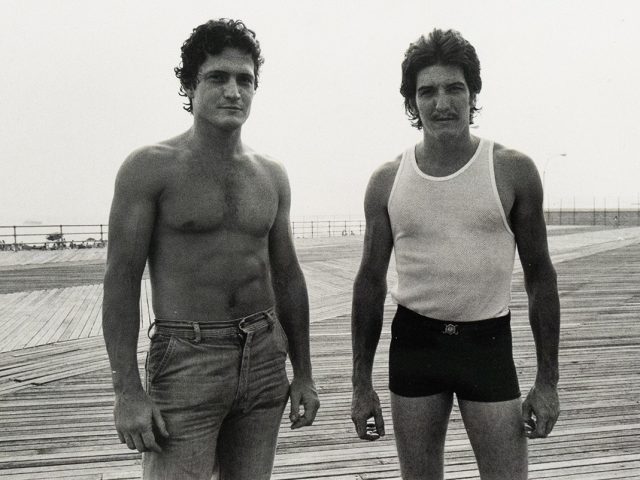Two men stand side-by-side on a wooden boardwalk, facing the viewer. The man on the left is shirtless and wears jeans, while the man on the right wears a tank top and dark shorts.