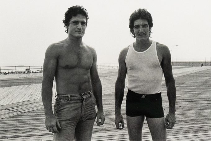 Two men stand side-by-side on a wooden boardwalk, facing the viewer. The man on the left is shirtless and wears jeans, while the man on the right wears a tank top and dark shorts.