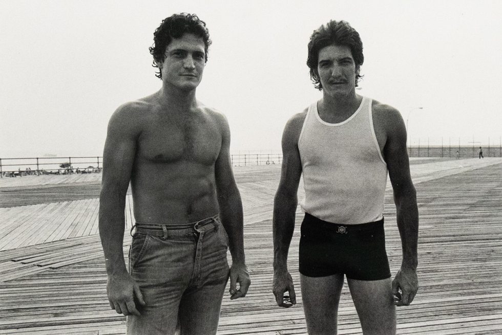 Two men stand side-by-side on a wooden boardwalk, facing the viewer. The man on the left is shirtless and wears jeans, while the man on the right wears a tank top and dark shorts.