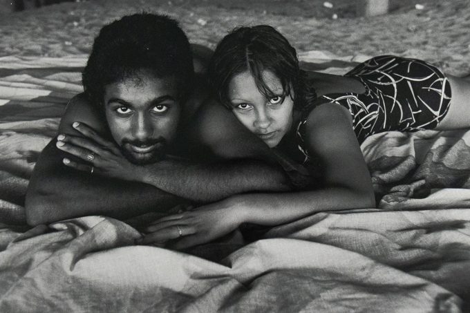 Black-and-white photograph of a man and woman lying side by side on a patterned blanket at the beach. Both are resting on their stomachs with their chins on their hands, gazing directly at the camera.