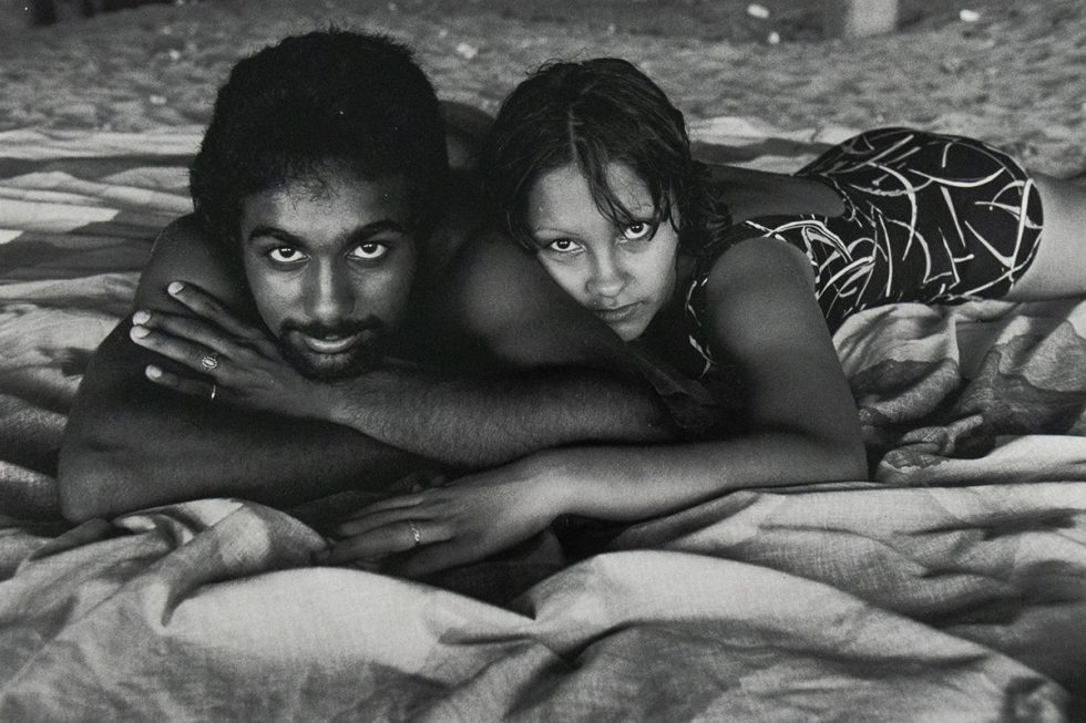 Black-and-white photograph of a man and woman lying side by side on a patterned blanket at the beach. Both are resting on their stomachs with their chins on their hands, gazing directly at the camera.