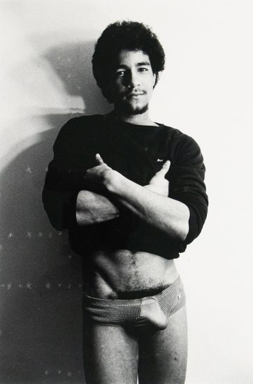 ChatGPT said: Black-and-white photograph of a man standing against a plain wall, wearing a dark long-sleeve shirt and patterned briefs. His arms are crossed over his chest, and he looks directly at the camera.