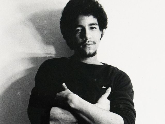 ChatGPT said: Black-and-white photograph of a man standing against a plain wall, wearing a dark long-sleeve shirt and patterned briefs. His arms are crossed over his chest, and he looks directly at the camera.