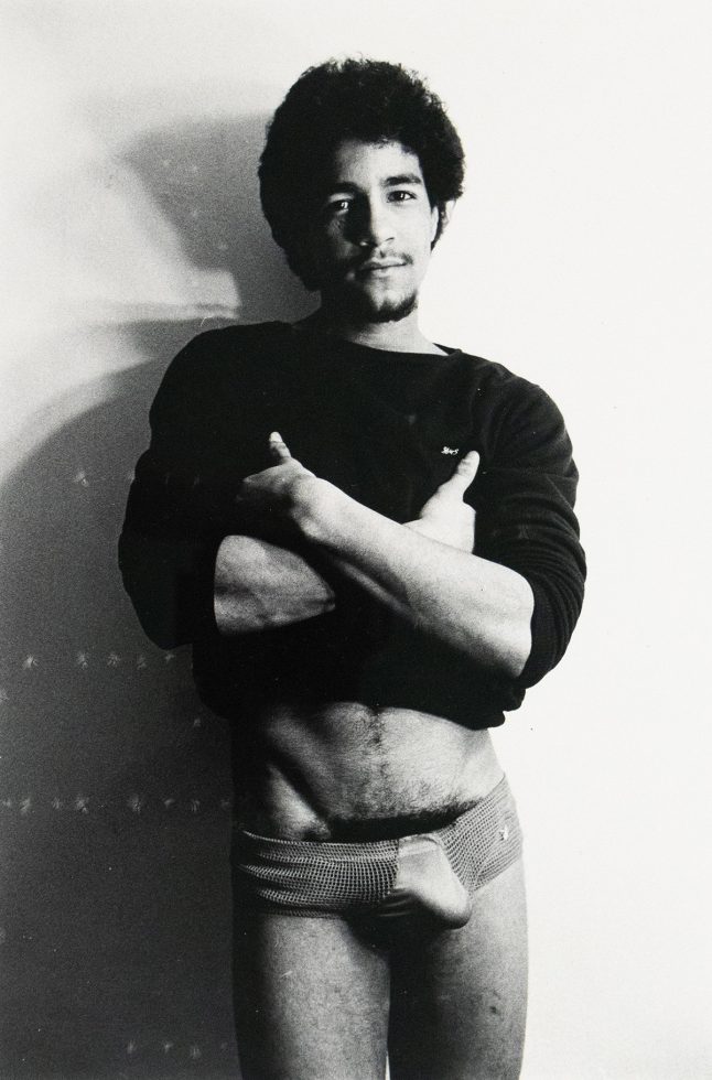 ChatGPT said: Black-and-white photograph of a man standing against a plain wall, wearing a dark long-sleeve shirt and patterned briefs. His arms are crossed over his chest, and he looks directly at the camera.