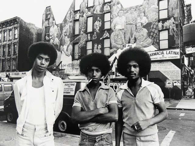 Three men with Afros stand on a city street, facing the viewer. Behind them, a large mural covers the side of a building, and a commercial street scene unfolds.