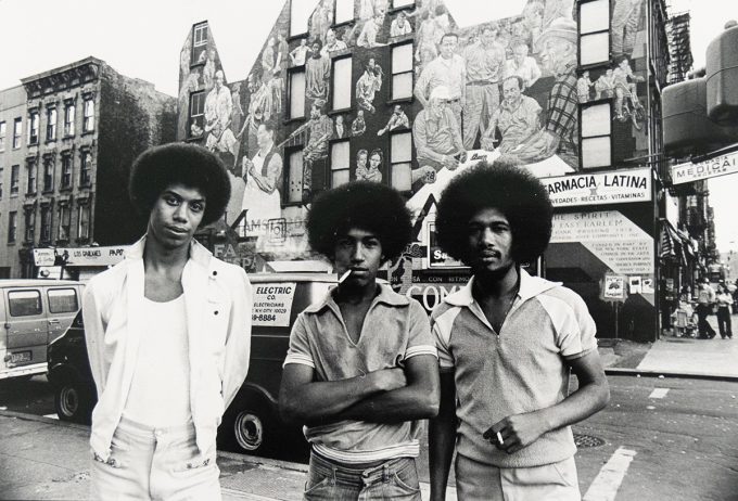 Three men with Afros stand on a city street, facing the viewer. Behind them, a large mural covers the side of a building, and a commercial street scene unfolds.