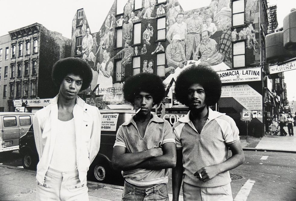 Three men with Afros stand on a city street, facing the viewer. Behind them, a large mural covers the side of a building, and a commercial street scene unfolds.