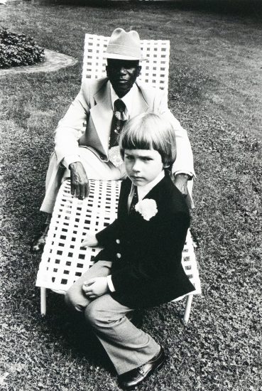 A black and white photograph captures an older man in a hat and suit sitting in a lawn chair, with a young child in a suit seated on his lap, looking directly at the viewer. They are outdoors on a grassy area.