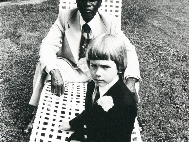 A black and white photograph captures an older man in a hat and suit sitting in a lawn chair, with a young child in a suit seated on his lap, looking directly at the viewer. They are outdoors on a grassy area.