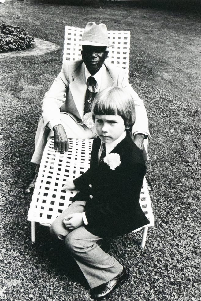A black and white photograph captures an older man in a hat and suit sitting in a lawn chair, with a young child in a suit seated on his lap, looking directly at the viewer. They are outdoors on a grassy area.