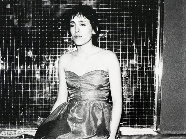 A black and white photograph shows a woman with short, dark hair sitting on a patterned bench in front of a grid-like reflective surface. She wears a strapless dress and fishnet stockings, with her legs crossed and her gaze directed forward.