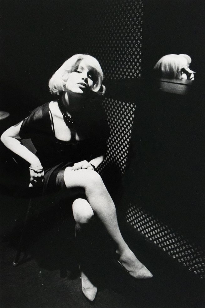 A blonde woman sits with crossed legs in a chair pushed up against a patterned wall. Her reflection is visible in a mirror behind her to the right.