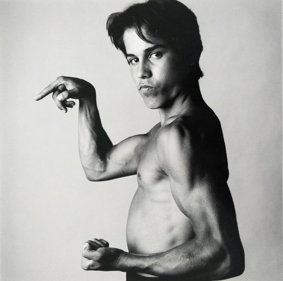 A shirtless young man poses in profile, flexing his arm muscles with a serious expression against a plain studio backdrop.