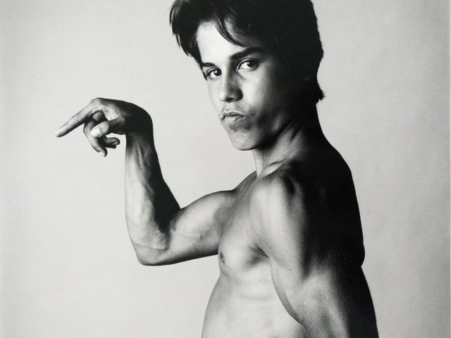 A shirtless young man poses in profile, flexing his arm muscles with a serious expression against a plain studio backdrop.