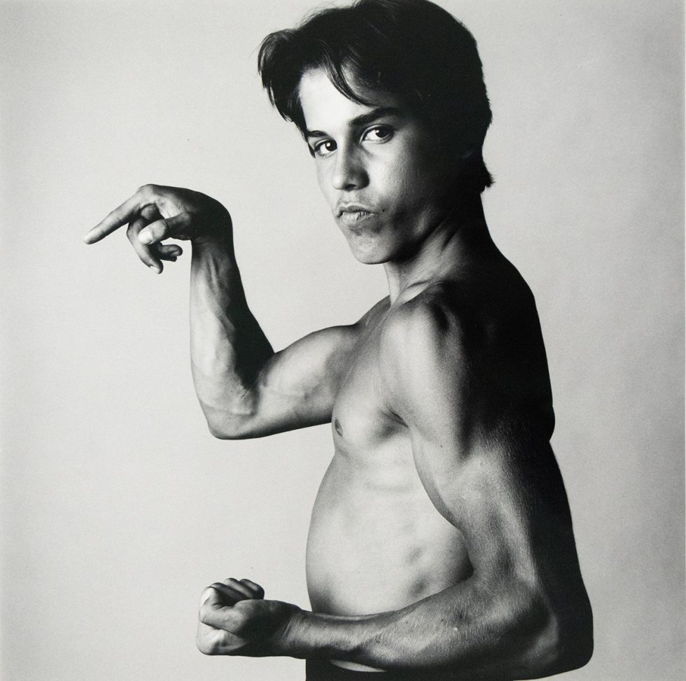 A shirtless young man poses in profile, flexing his arm muscles with a serious expression against a plain studio backdrop.