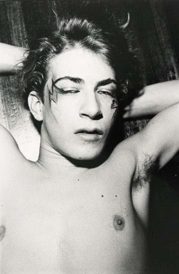A shirtless man with lightning bolt-shaped face paint lies on his back with eyes closed, hands behind his head. The black and white image shows his chest and face in close-up.
