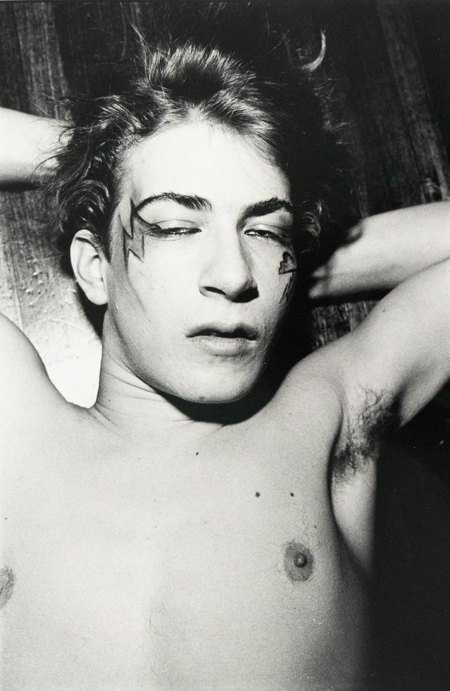 A shirtless man with lightning bolt-shaped face paint lies on his back with eyes closed, hands behind his head. The black and white image shows his chest and face in close-up.