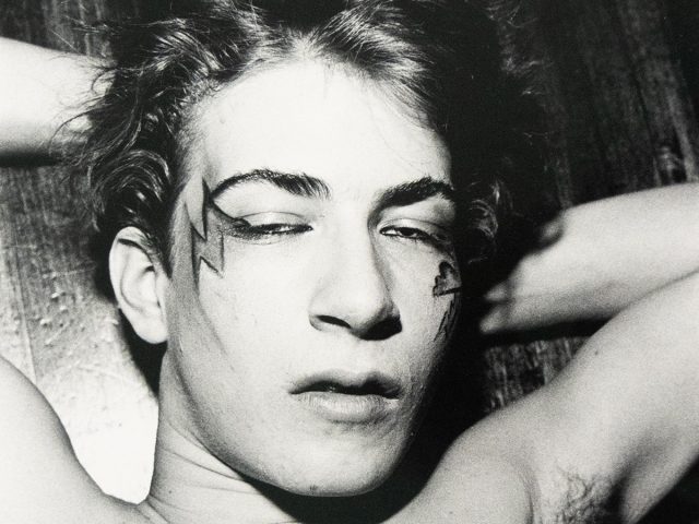 A shirtless man with lightning bolt-shaped face paint lies on his back with eyes closed, hands behind his head. The black and white image shows his chest and face in close-up.