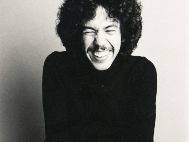 A black and white studio portrait captures a person with curly hair and a mustache, seated and smiling broadly, wearing a dark turtleneck and dark pants. They are looking directly at the viewer with visible joy.